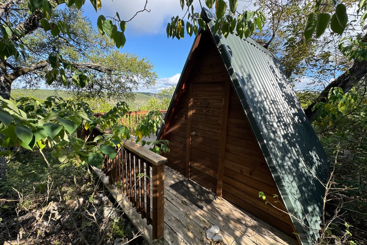 A wooden A-frame glamping cabin with a green metal roof, tucked into the trees beside its deck.
