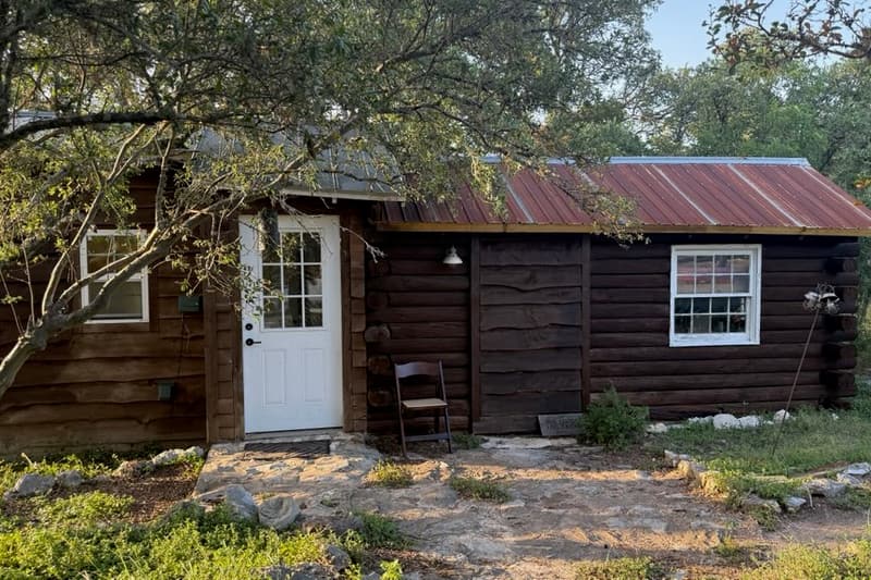The Little Log Cabin: dark logs, a white door, and a rust-red tin roof set back among the trees.