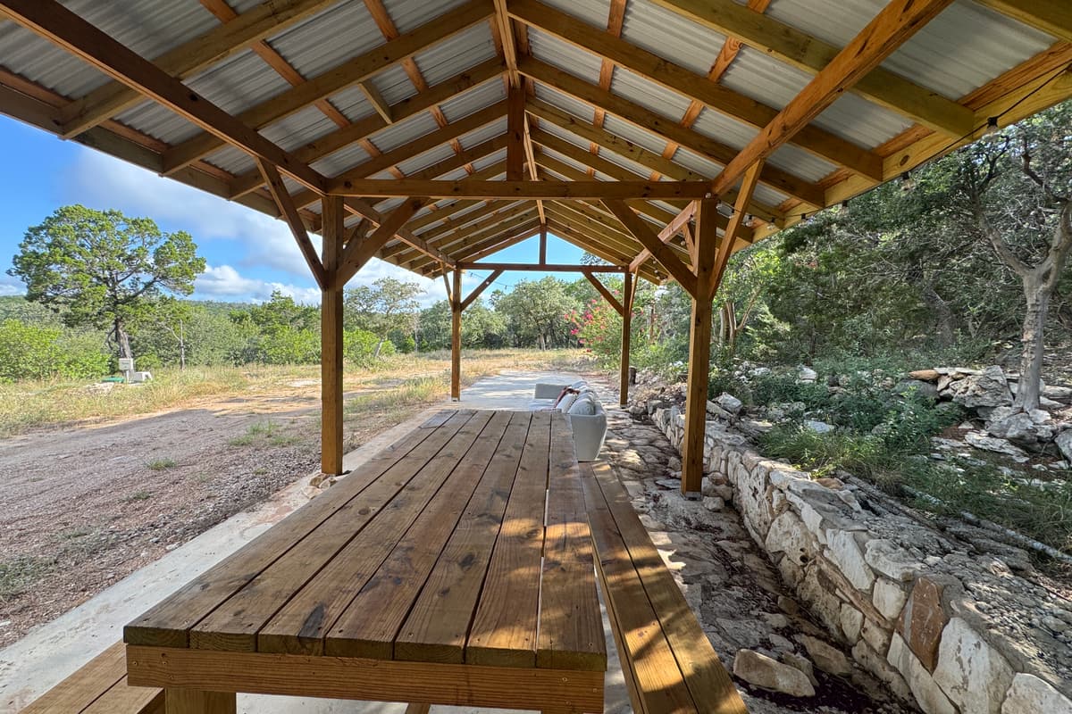 An open-air pavilion with a long wooden table and benches, looking out over trees and hills.
