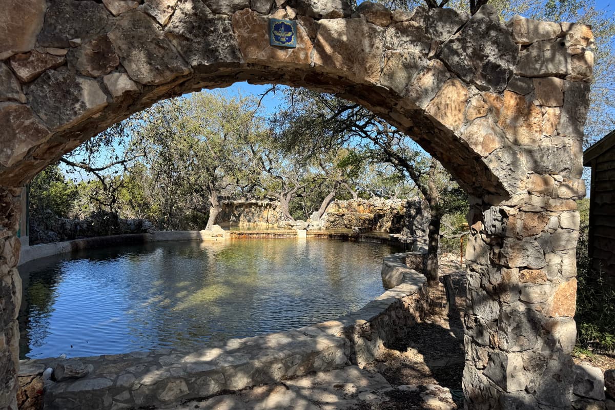 A hand-built stone arch framing the natural stone pool beneath old live oaks.